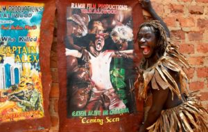 In this photo taken Wednesday, Sept. 7, 2016, actor Francis Kagoro pulls a face for the camera while showing posters of previous movies, at the "Wakaliwood" studios in the Wakaliga slum of Kampala, Uganda. Deep in this Kampala slum at a tin-roofed collection of houses known as Wakaliwood, is the engine of Uganda's tiny film industry and the source of $200-budget movies and a glimmer of fame. (AP Photo/Stephen Wandera)