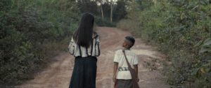 A young woman and boy walk along the path together, facing away from the camera, in Mattie Do's THE LONG WALK