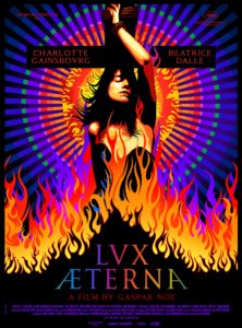 Charlotte Gainsbourg poses against a cross while flames engulf her up to the chest against a vivid red, yellow, and purple color background